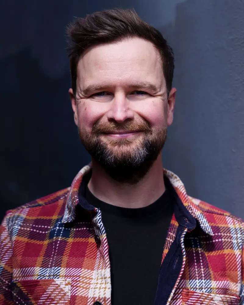 Alan in a red checked overshirt, smiling, against a dark wall