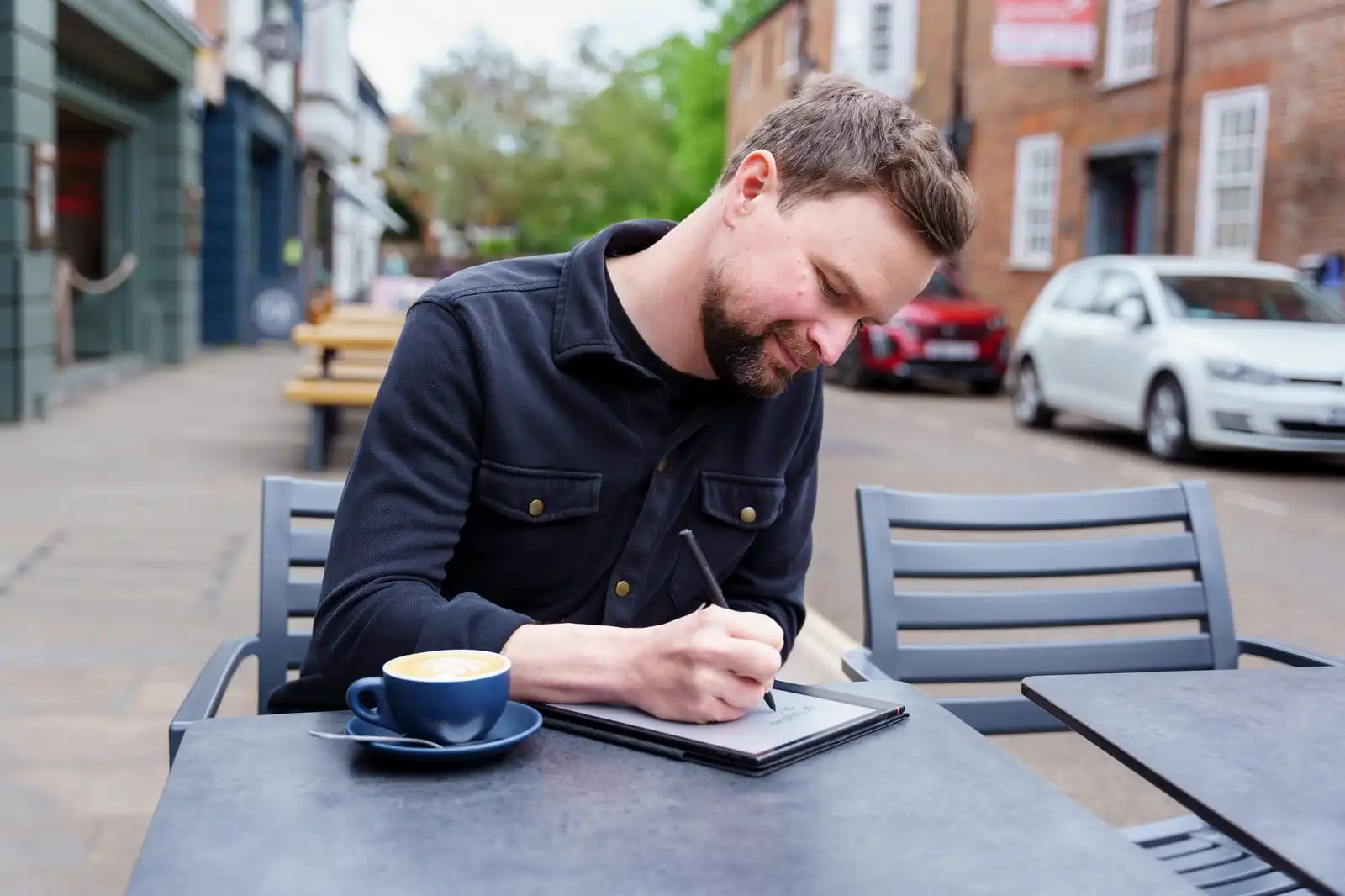 Alan writing at a café table outdoors, coffee beside him